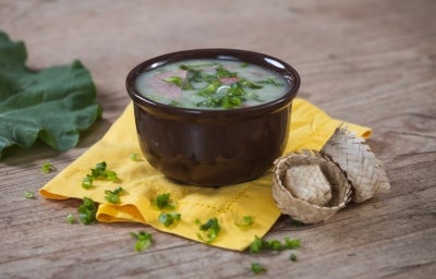 Imagem de um bowl marrom com a receita de caldo verde polvilhada com cebolinha sobre um pano amarelo e chapéus de festa junina ao lado.