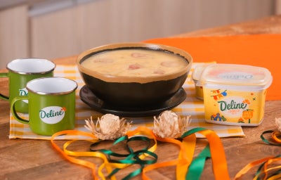 Caldo cremoso de macaxeira com rodelas de calabresa em tigela, acompanhado de manteiga Deline e canecas para festa junina