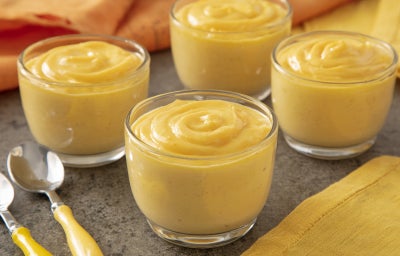 Imagem mostra 4 copinhos transparentes com um creme amarelo dentro dispostos sob uma superfície cinza e amarela com colheres