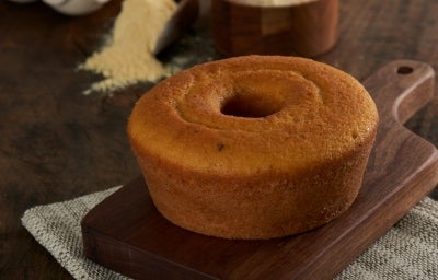 Foto aproximada do bolo de milho com erva-doce, redondo, com furo central, bem dourado, sobre uma tábua de madeira com um punhado de fubá aparecendo ao fundo
